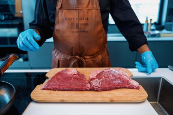 Två bitar rå flankstek på en skärbräda framför en kock i Meat District-förkläde
